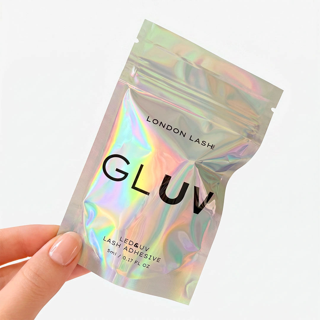 A bottle of London Lash Gluv LED & UV Lash Adhesive with a metallic purple cap and a dark gray/black base, shown on a clean white background with a few clear adhesive drops in the foreground.