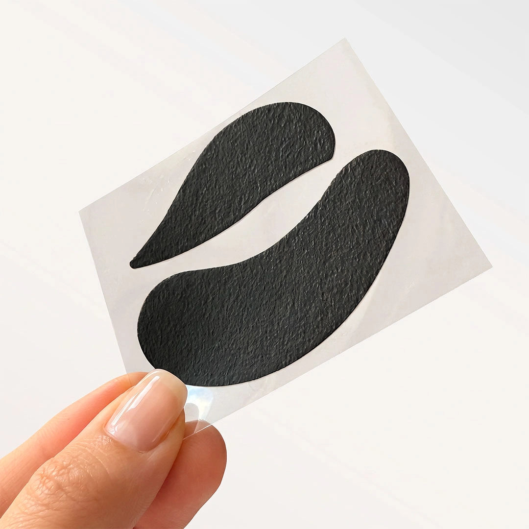 A hand holding a small white sheet with two black teardrop-shaped eye patches against a clean white background.