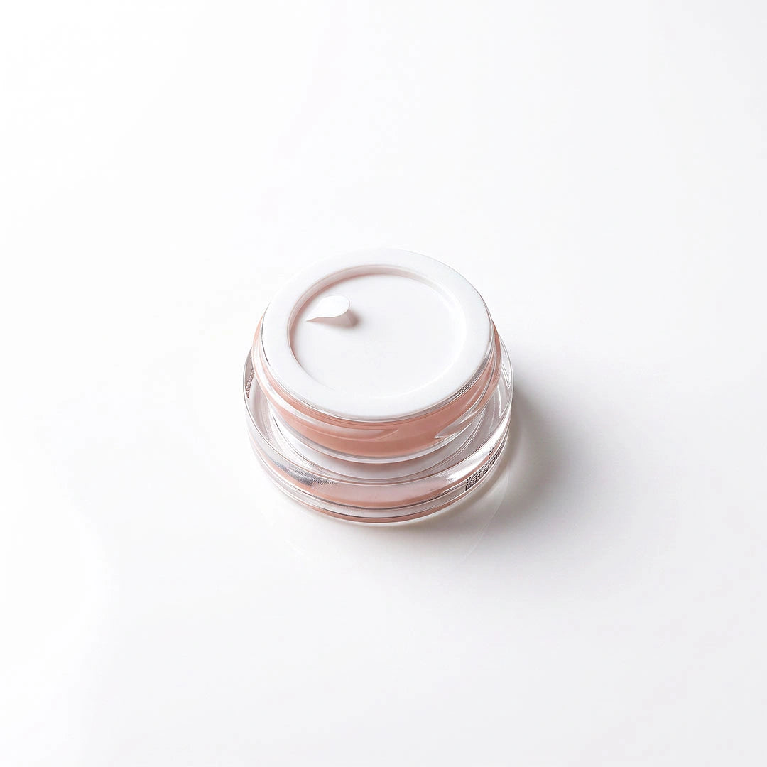A small clear cosmetic jar with a white lid, containing a pale peach-pink cream, photographed on a clean white background.