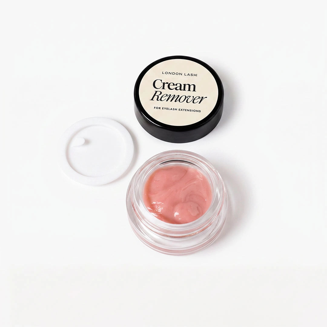 A small clear cosmetic jar containing pink cream remover, shown open with its black lid placed behind it on a clean white background. The lid label reads 'London Lash Cream Remover for Eyelash Extensions.