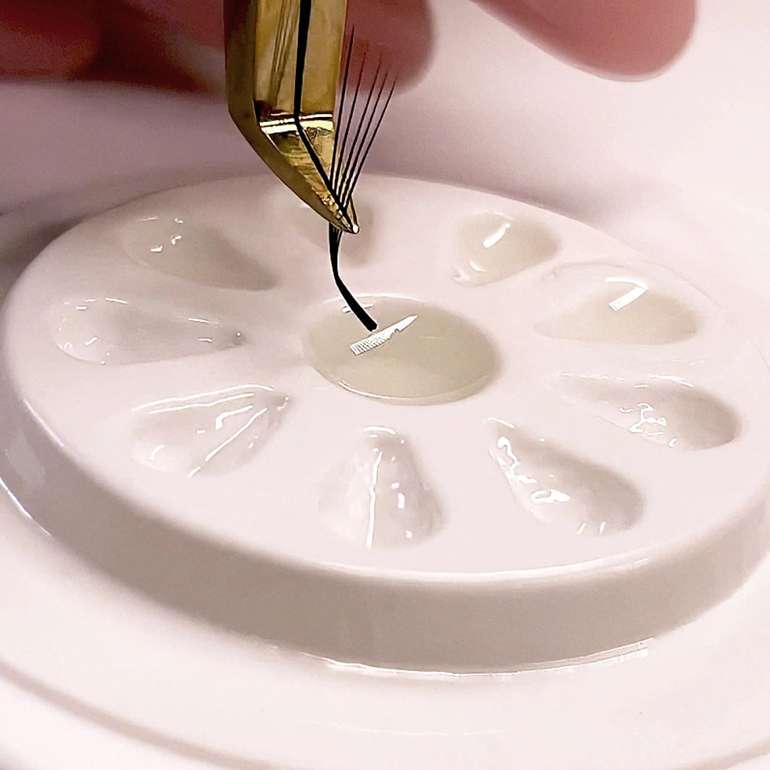 A close-up of eyelash extension fan being held by tweezers above a small round well of adhesive on a white petri-dish-like surface.