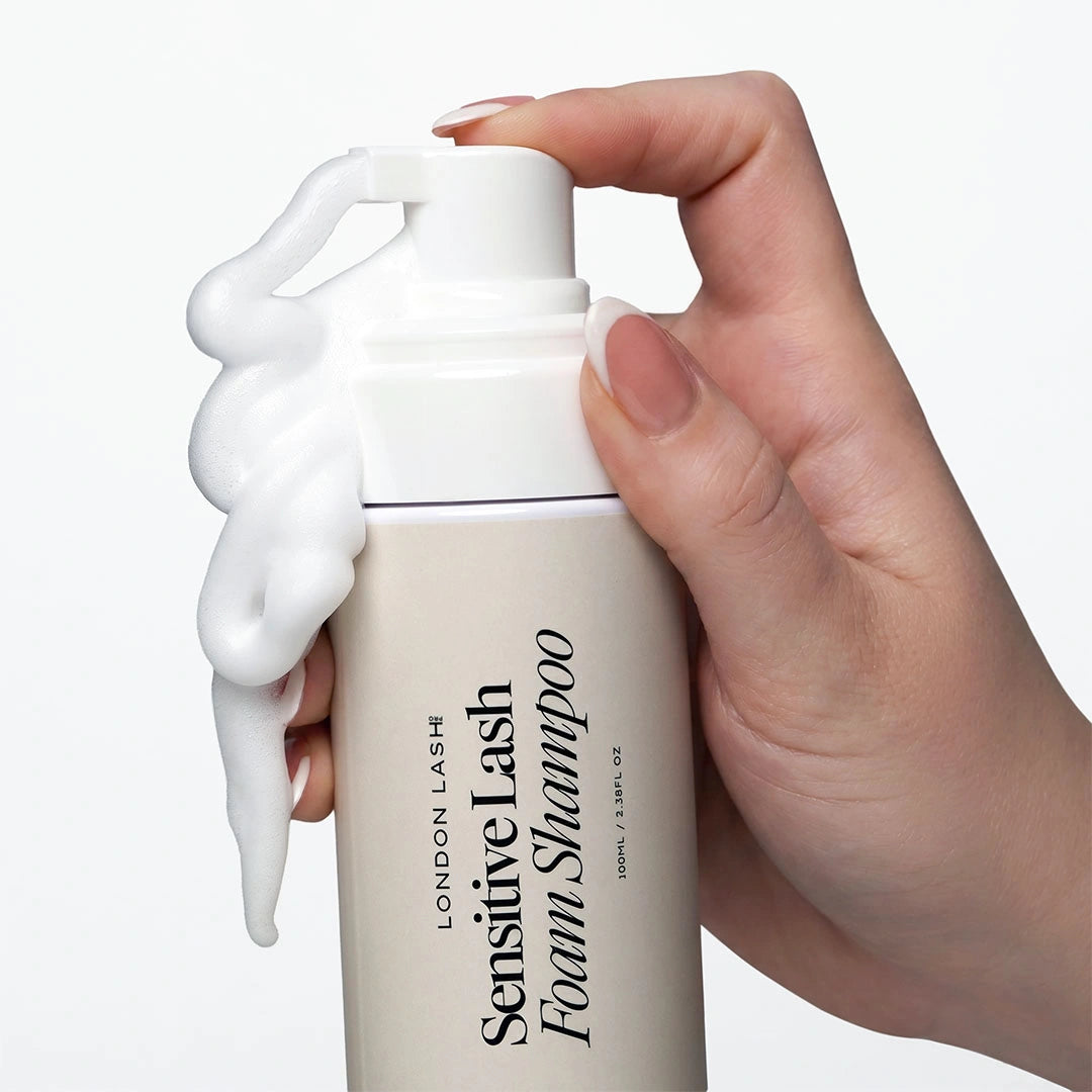A close-up shot of a hand with a French manicure pressing down on the pump of the lash shampoo bottle, causing thick, white foam to spill down the side of the container