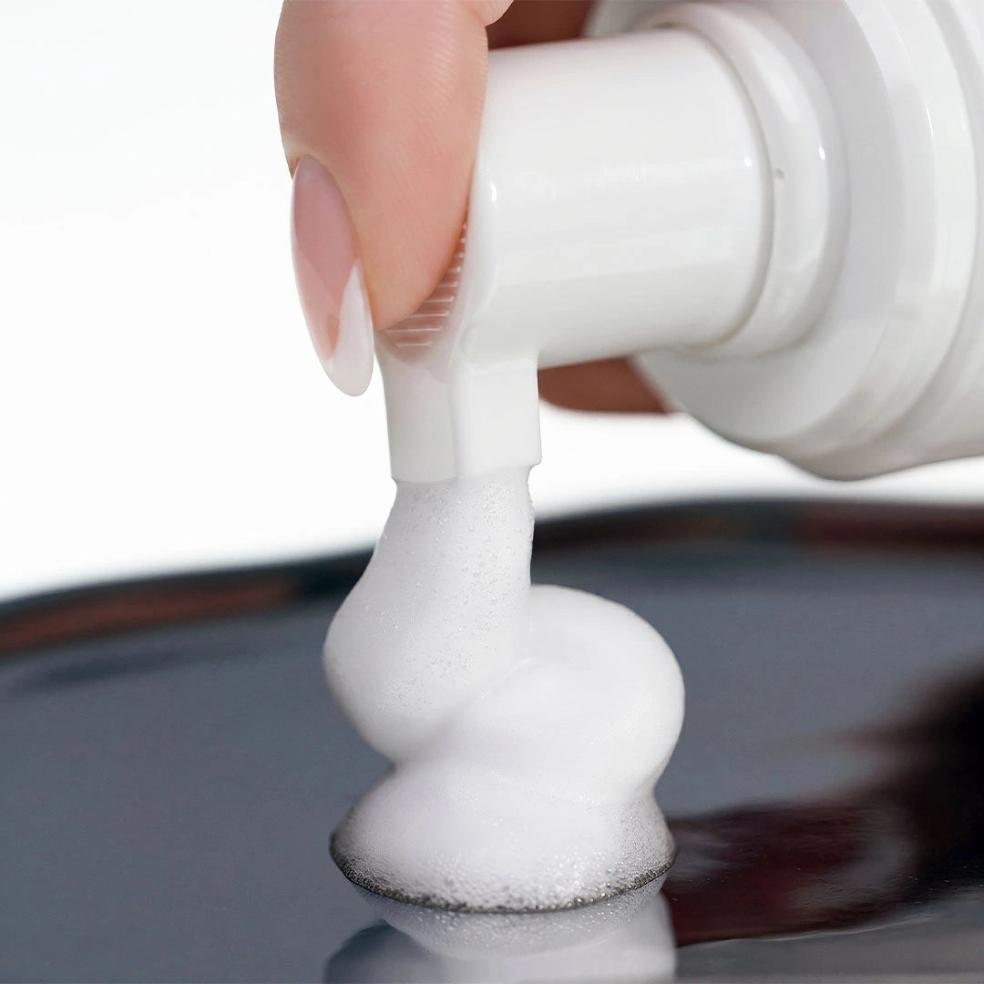 Close-up view of a finger pressing the nozzle of a white pump bottle, dispensing a thick, smooth dollop of white foaming shampoo onto a dark, reflective metallic surface.