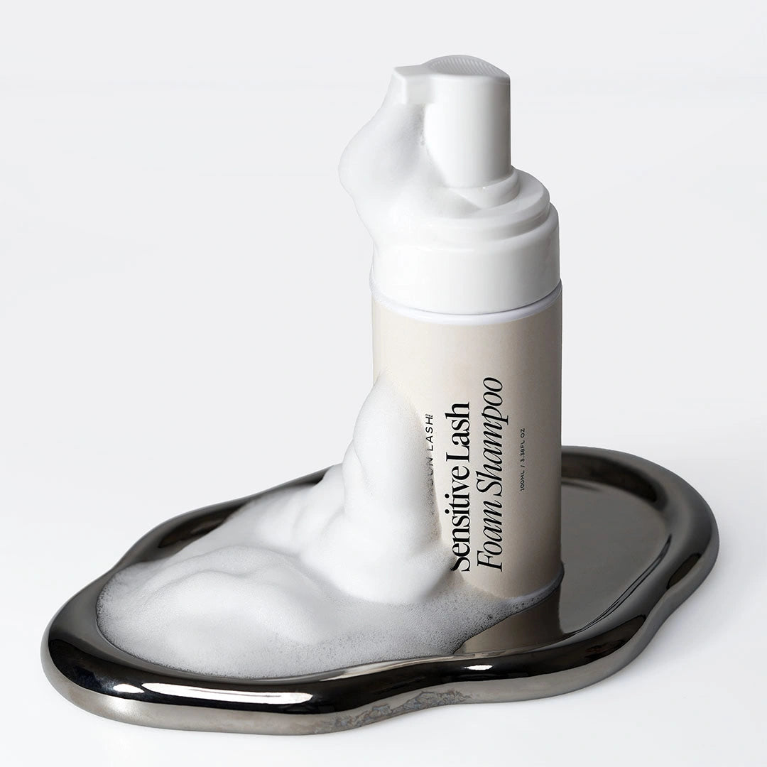 A white bottle of London Lash Sensitive Lash Foam Shampoo standing on a wavy, reflective silver tray, with a large, artistic spill of thick white foam cascading down the bottle and pooling onto the tray.